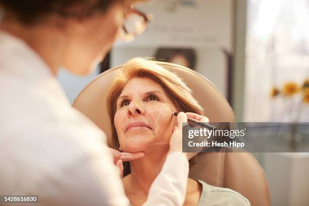 female doctor drawing correction lines on woman's face - chirurgo plastico foto e immagini stock