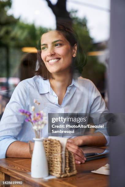 portrait shot through window of smiling latin-american woman sitting at cafe - mature latin women stockfoto's en -beelden