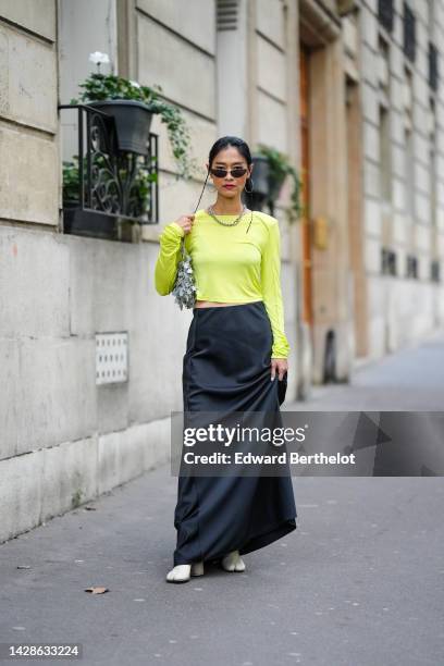 Pornwika Spiecker wears earrings, sunglasses, a necklace, a neon yellow top, a black maxi silky lustrous skirt, white shoes with separate toes, a...