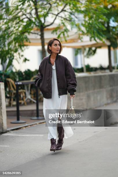 Geraldine Boublil wears a brown bomber jacket, a white slit maxi dress, brown pointed boots, a white bag, outside Acne, during Paris Fashion Week -...