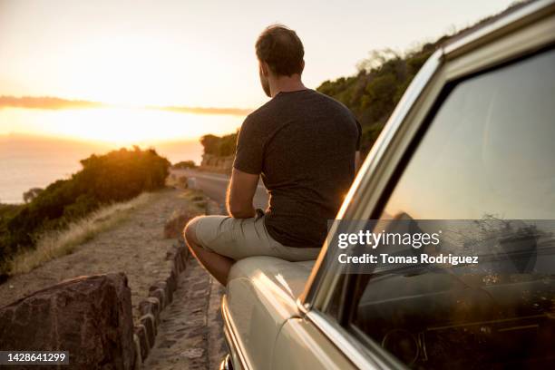 man sitting on car hood at coastal road looking at view - car hood stock pictures, royalty-free photos & images