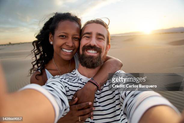cheerful selfie of lovely multiracial just married couple at desert with a beautiful sunset background. - couple dunes stockfoto's en -beelden