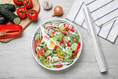 Bowl of fresh salad with plastic food wrap on white wooden table, flat lay