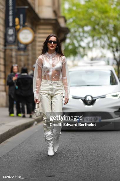 Mary Leest wears brown sunglasses, gold earrings, a white transparent shirt, a white bra underwear, white shiny leather large pants, a white shiny...