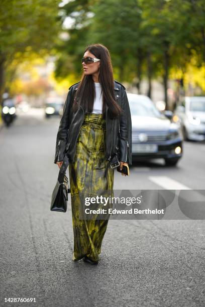 Guest wears sunglasses, a white t-shirt, a black shiny leather oversized biker jacket, a black and yellow shiny leather tie and dye print pattern...