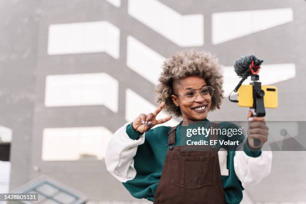 trendy african american woman influencer filming video with smartphone doing peace victory sign with hand - american influencer - fotografias e filmes do acervo