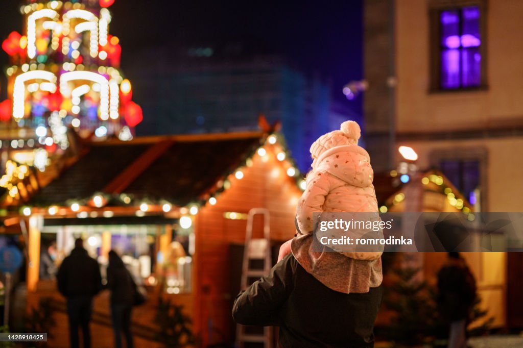 Petite fille d’âge préscolaire assise sur l’épaule de son père sur le marché de Noël en Allemagne. Enfant en bas âge heureux et homme observant la pyramide décorée traditionnelle. Famille heureuse, liens, amour. Noël en famille.