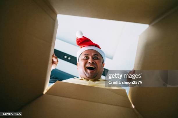 excited asian man wearing santa hat unpacking christmas gift. - christmas surprise stock pictures, royalty-free photos & images