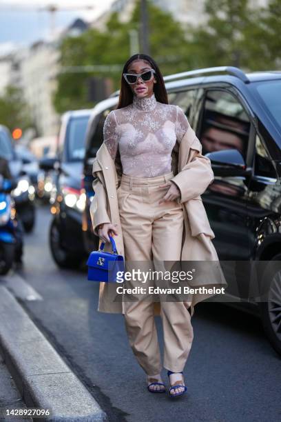 Winnie Harlow wears gray marble print pattern butterfly sunglasses, a white lace print pattern turtleneck / long sleeves t-shirt, a beige long trench...