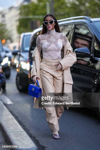 Winnie Harlow wears gray marble print pattern butterfly sunglasses, a white lace print pattern turtleneck / long sleeves t-shirt, a beige long trench...