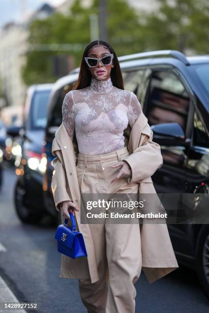 Winnie Harlow wears gray marble print pattern butterfly sunglasses, a white lace print pattern turtleneck / long sleeves t-shirt, a beige long trench...