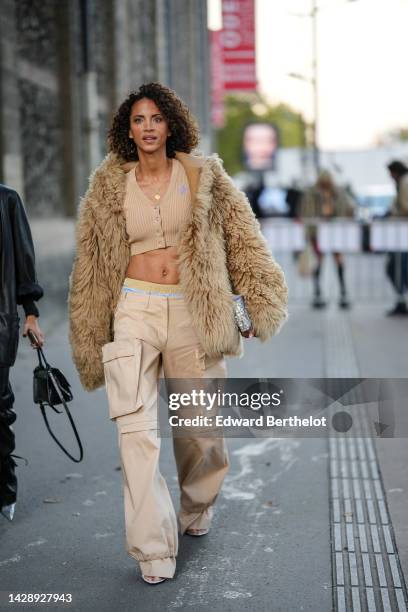 Noemie Lenoir wears a gold long chain necklace, a brown ribbed wool buttoned / V-neck / cropped cardigan, a brown fluffy oversized hoodie coat, beige...
