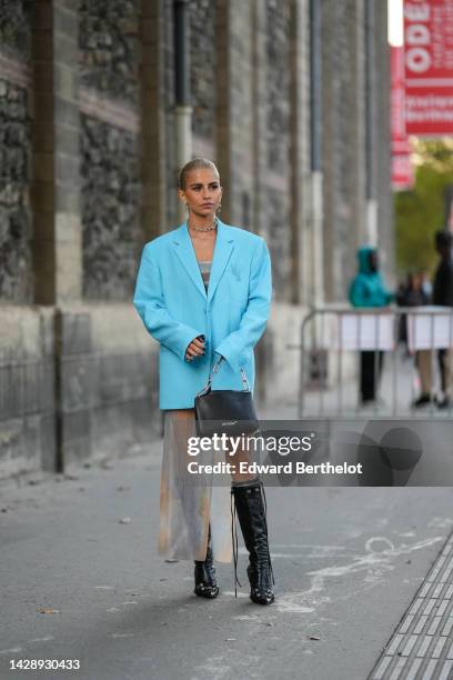 Caroline Daur wears gold large earrings, a matching silver large chain necklace, a blue oversized blazer jacket, a white ribbed wool asymmetric long...