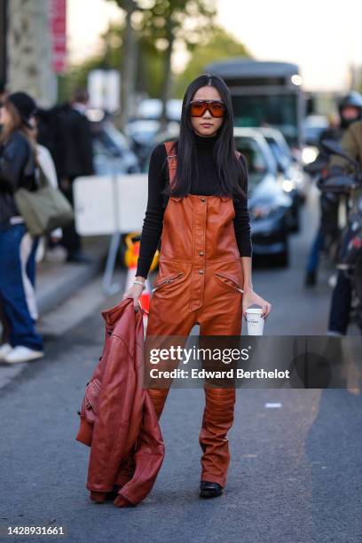 Yoyo Cao wears brown and orange sunglasses, a black turtleneck / long sleeves wool pullover, camel shiny leather buttoned / zipper overalls, a brown...