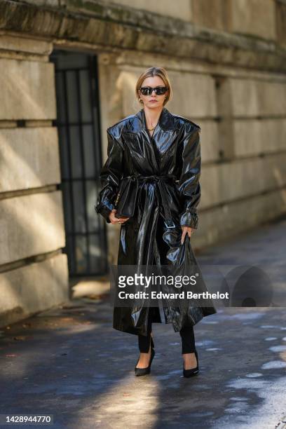 Elizabeth Sulcer wears black sunglasses, gold earrings, a black shiny varnished leather oversized / belted long dress, black legging pants, a black...