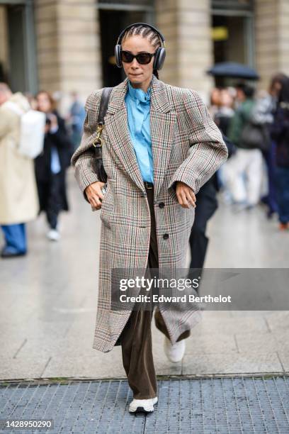 Adwoa Aboah wears a pale blue high neck shirt, a brown / red / beige houndstooth print pattern long coat, black headphones, brown ribbed velvet...