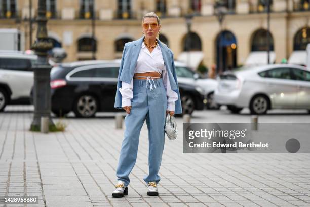 Olga Ferrara wears orange sunglasses, gold earrings, a gold large chain necklace, a white laces waist / cropped shirt, a pale blue cropped denim...