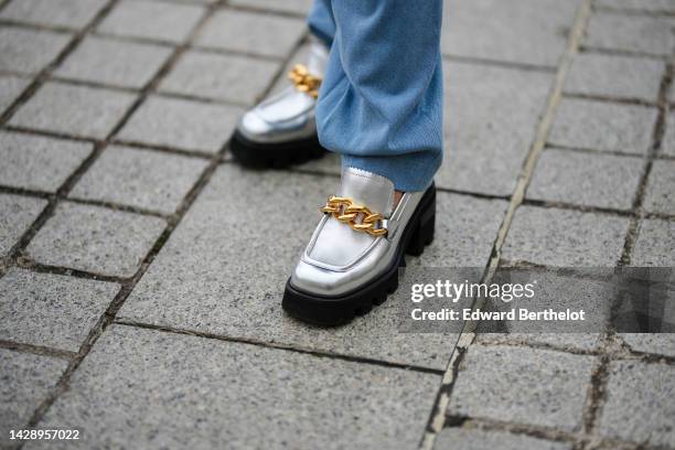 Olga Ferrara wears pale blue denim large pants, silver shiny lather with embroidered large gold chain toe-cap / block heels loafers, outside Chloe,...