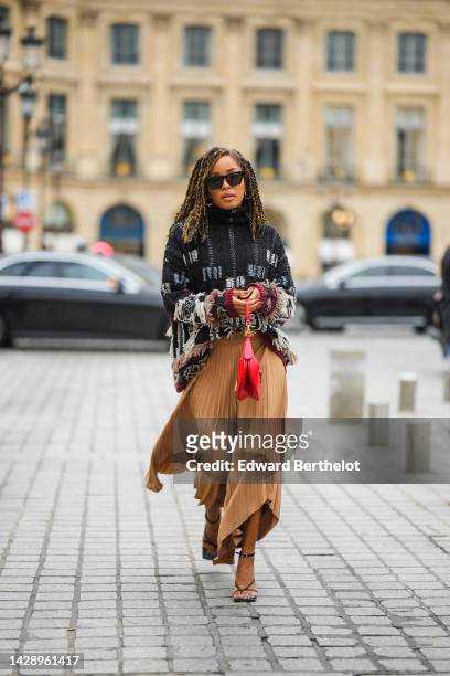 Ellie Delphine wears black sunglasses, gold earrings, a black with embroidered white and burgundy ripped wool pattern high neck / pullover, a brown...