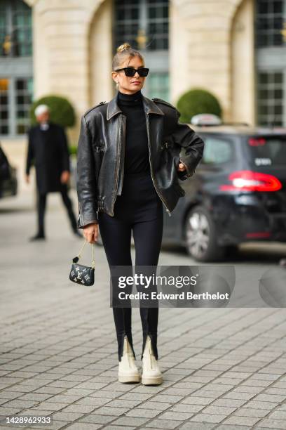 Xenia Adonts wears black sunglasses, white pearls earrings, a black turtleneck pullover from Prada, a black shiny leather zipper oversized jacket,...