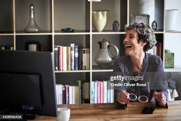 laughing mature woman sitting at table with  desktop pc at home - freelance work candid stock pictures, royalty-free photos & images