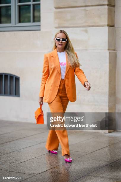 Guest wears white square sunglasses, diamonds earrings, a white with neon pink inscriptions print pattern t-shirt, an orange blazer jacket, matching...