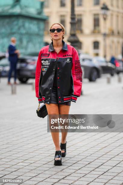 Guest wears black square sunglasses, gold earrings, a red and black shiny leather with embroidered inscriptions print pattern bomber coat, a black...