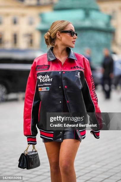 Guest wears black square sunglasses, gold earrings, a red and black shiny leather with embroidered inscriptions print pattern bomber coat, a black...