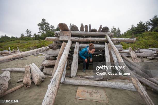 a young woman exits a beach shelter made entirely out of driftwood on wickaninnish beach - pacific rim national park reserve stock pictures, royalty-free photos & images