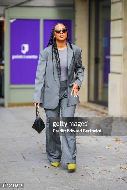 Guest wears black sunglasses, a gray t-shirt, a gray oversized blazer jacket, matching gray flared suit pants, a black shiny leather triangle shape...