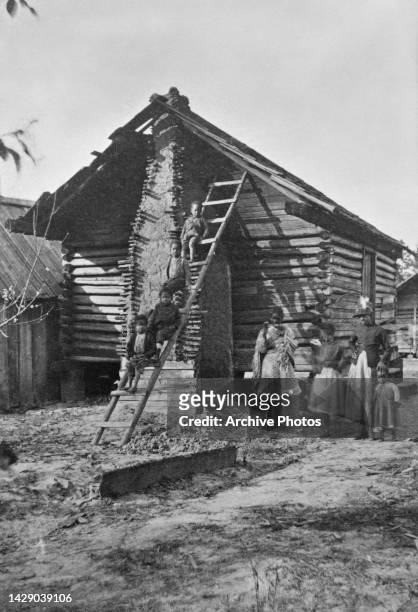 Four children sitting on a ladder which leans against a log cabin, their family's quarters, with young women and girls standing beneath the ladder,...