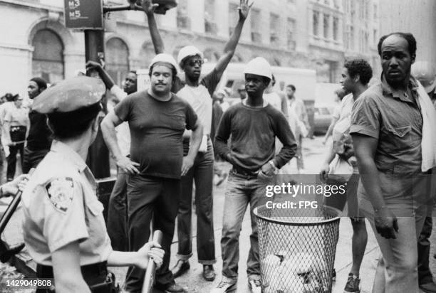 An NYPD police officer holding their nightstick as they approach a group of men, some wearing hardhats, on an in New York City, New York, circa 1980.