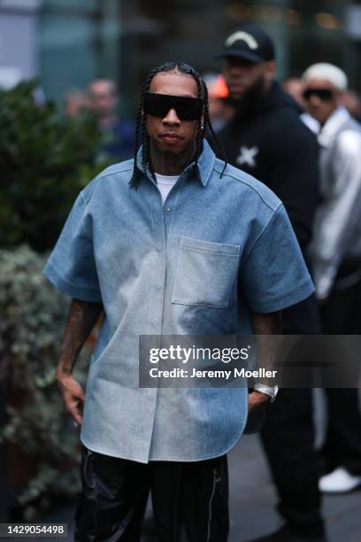 Tyga is seen wearing black futurist sunglasses, a white t-shirt, blue and white faded denim oversized short sleeves shirt and a black leather pants,...