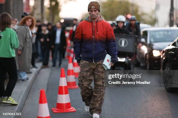 Marc Forne is seen wearing a brown Carhartt beanie, a red and blue faded Off-White bomber jacket, green camouflage pattern military cargo pants,...