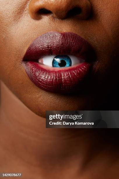 a dark-skinned african-american girl holds a plastic blue eye in her lips. large lips with a beautiful dark lipstick. beauty art concept of surrealism with copy space. - magic eye fotografías e imágenes de stock