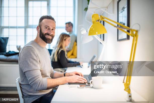 mature programmer working on laptop in co-working space. bearded employee smiling and looking at camera - code 41 stock pictures, royalty-free photos & images