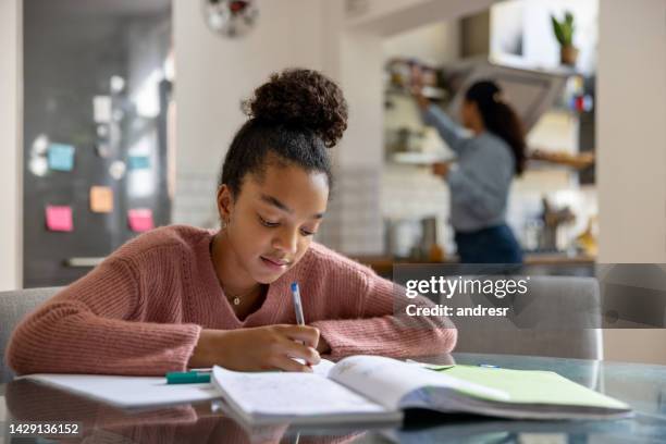 adolescente haciendo su tarea mientras su madre está cocinando en el fondo - ensayo fotografías e imágenes de stock