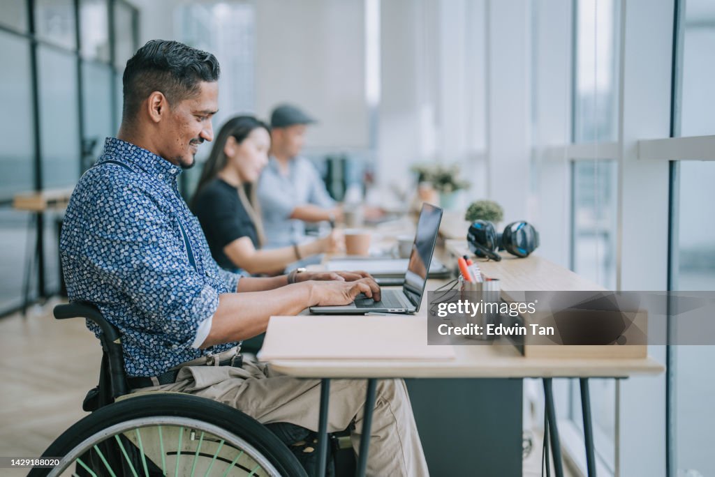 Asian Indian white collar male worker in wheelchair concentrating working in office beside his colleague