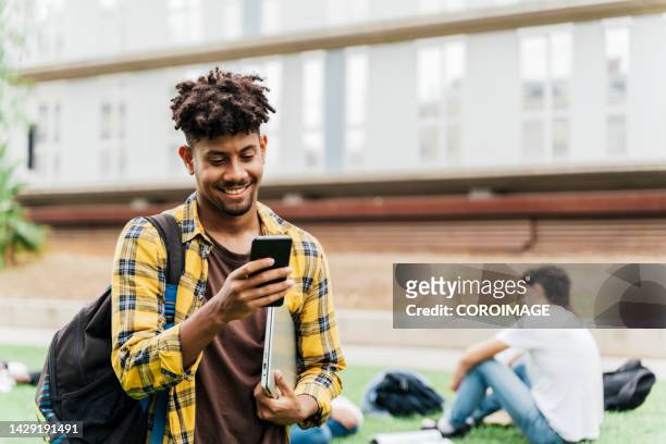 college student on college campus walking and looking at smart phone holding laptop. concept of students and technology. - young men walking stock pictures, royalty-free photos & images