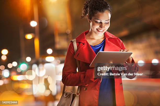 woman looking at tablet computer standing in city at night. - blue coat stock-fotos und bilder