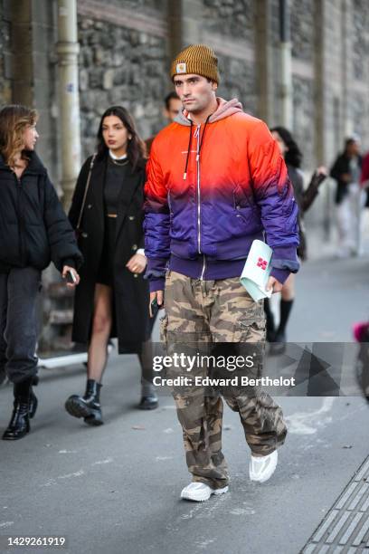 Guest wears a brown ribbed wool beanie from Carhartt, a white t-shirt, a gray and orange tie and dye print pattern hoodie sweater, a neon orange and...