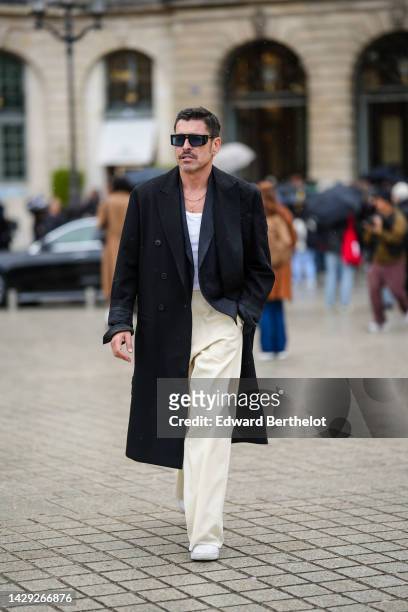 Alex Badia wears black sunglasses, a gold pendant cross earring, gold chain necklaces, a black embroidered flower pattern coat, a white tank-top, a...