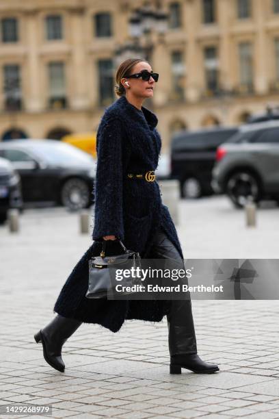 Guest wears black sunglasses from Prada, gold earrings, a black wool pullover, a navy blue and black braided wool long coat, a black shiny leather...