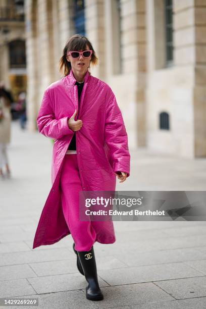 Guest wears pink sunglasses, a white t-shirt, a black wool buttoned cardigan, a neon pink shiny quilted long coat, neon pink large sport pants, black...