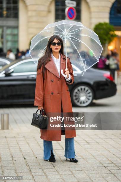 Guest wears a white transparent umbrella, a white shirt, a brown shiny leather long coat, blue denim ripped flared pants, a black shiny leather...