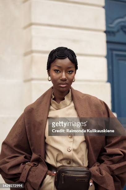 Guest wearing a light beige top and shorts, dark brown belt bag, long brown trench coat, and golden sandals and having braided hair in circles...