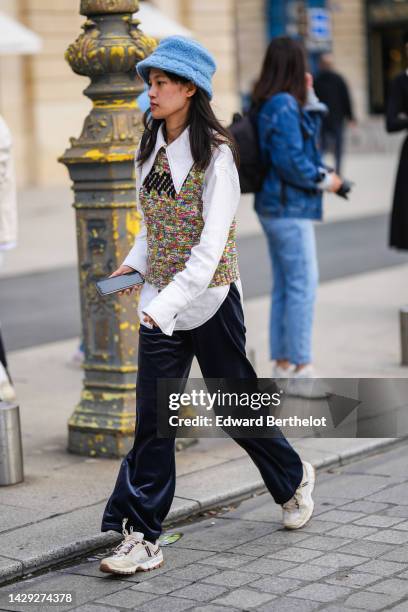 Guest wears a pale blue fluffy bob, a white shirt with a large collar, a multicolored braided wool sleeveless pullover, navy blue velvet large pants,...