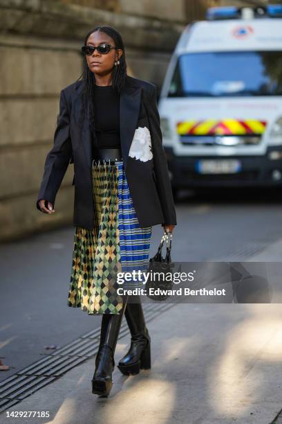 Guest wears black sunglasses, silver earrings, a black blazer jacket, a black t-shirt, a black shiny leather belt, a yellow / black / green...