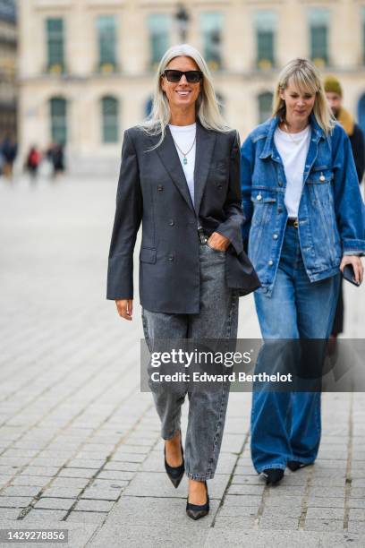 Sarah Harris wears black sunglasses, a white t-shirt, a gold pendant necklace, a dark gray blazer jacket, gray denim large pants, black nylon and...