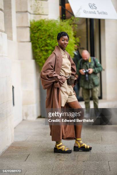 Guest wears gold earrings, a beige high neck buttoned short jumpsuit, a brown long oversized coat, a brown shiny leather belt bag, gold socks, gold...